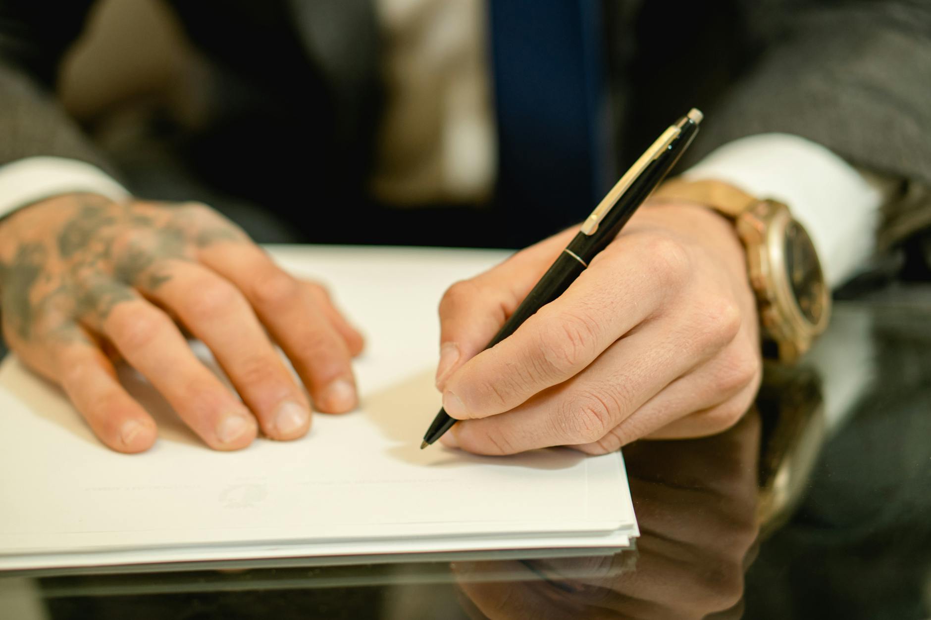 A close-up view of a businessman with tattoos signing a document at a desk.