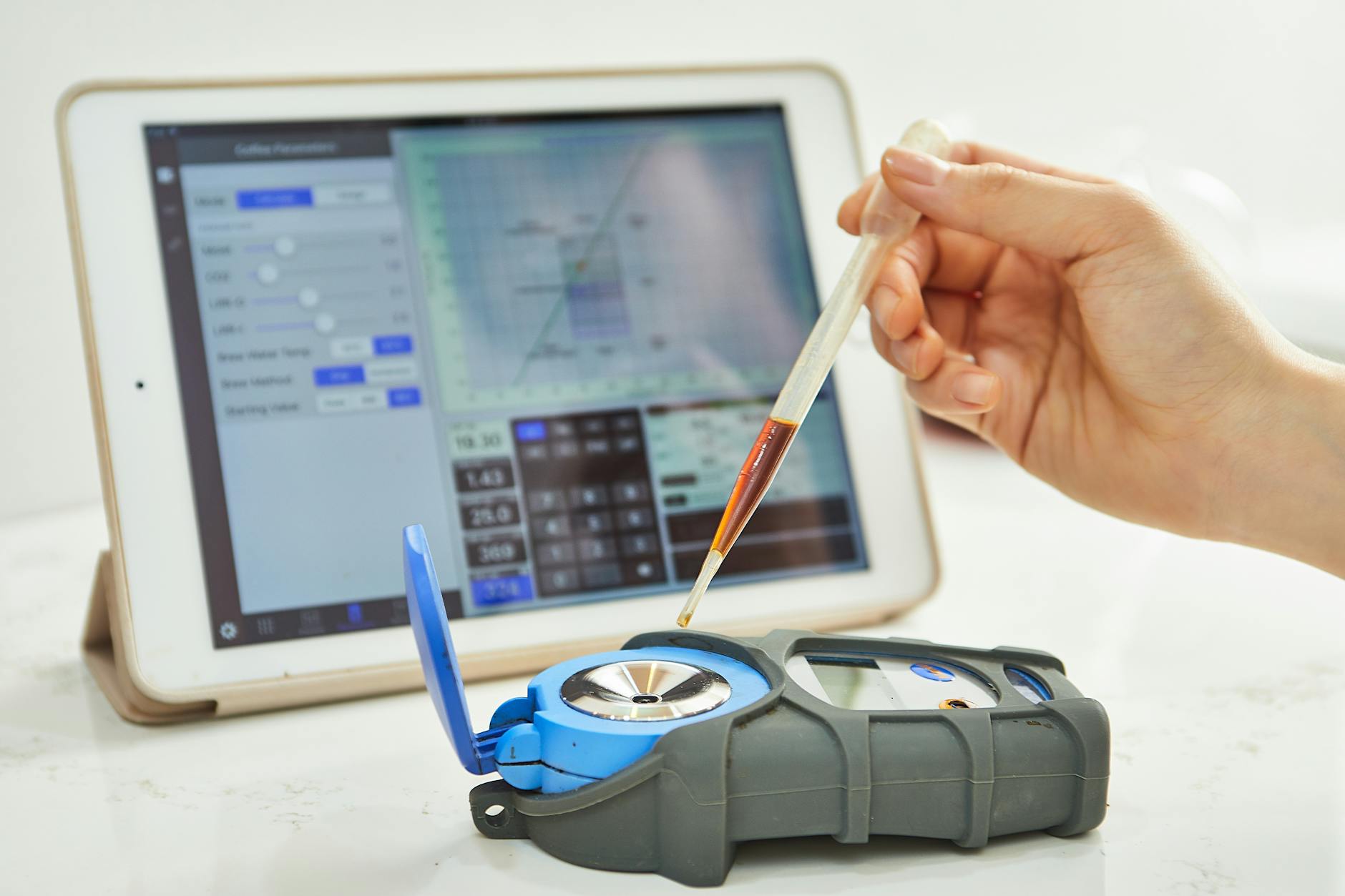 Hand using pipette with refractometer in front of tablet displaying measurement interface.