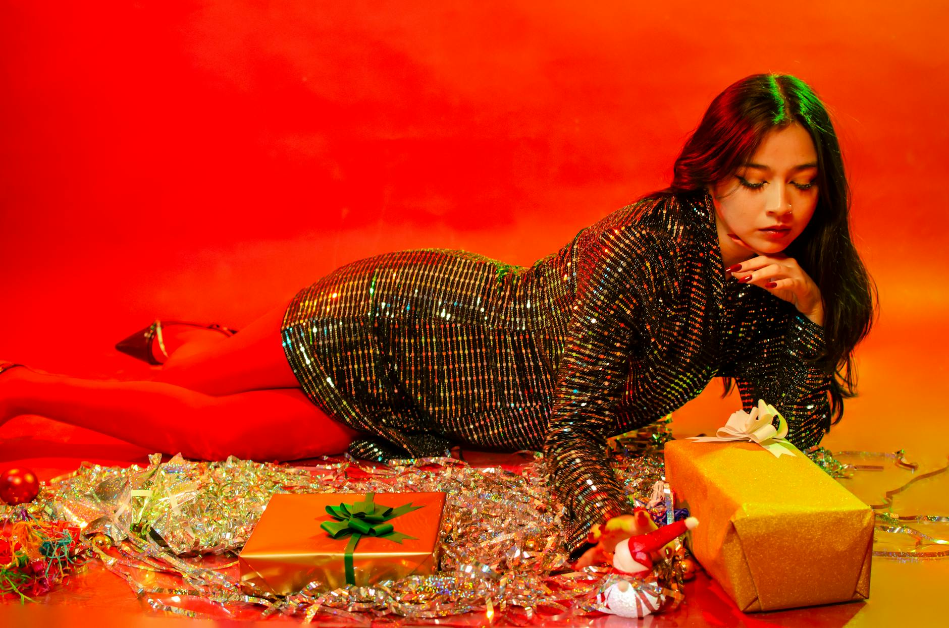 Model in shimmering outfit surrounded by Christmas gifts, exuding festive elegance.