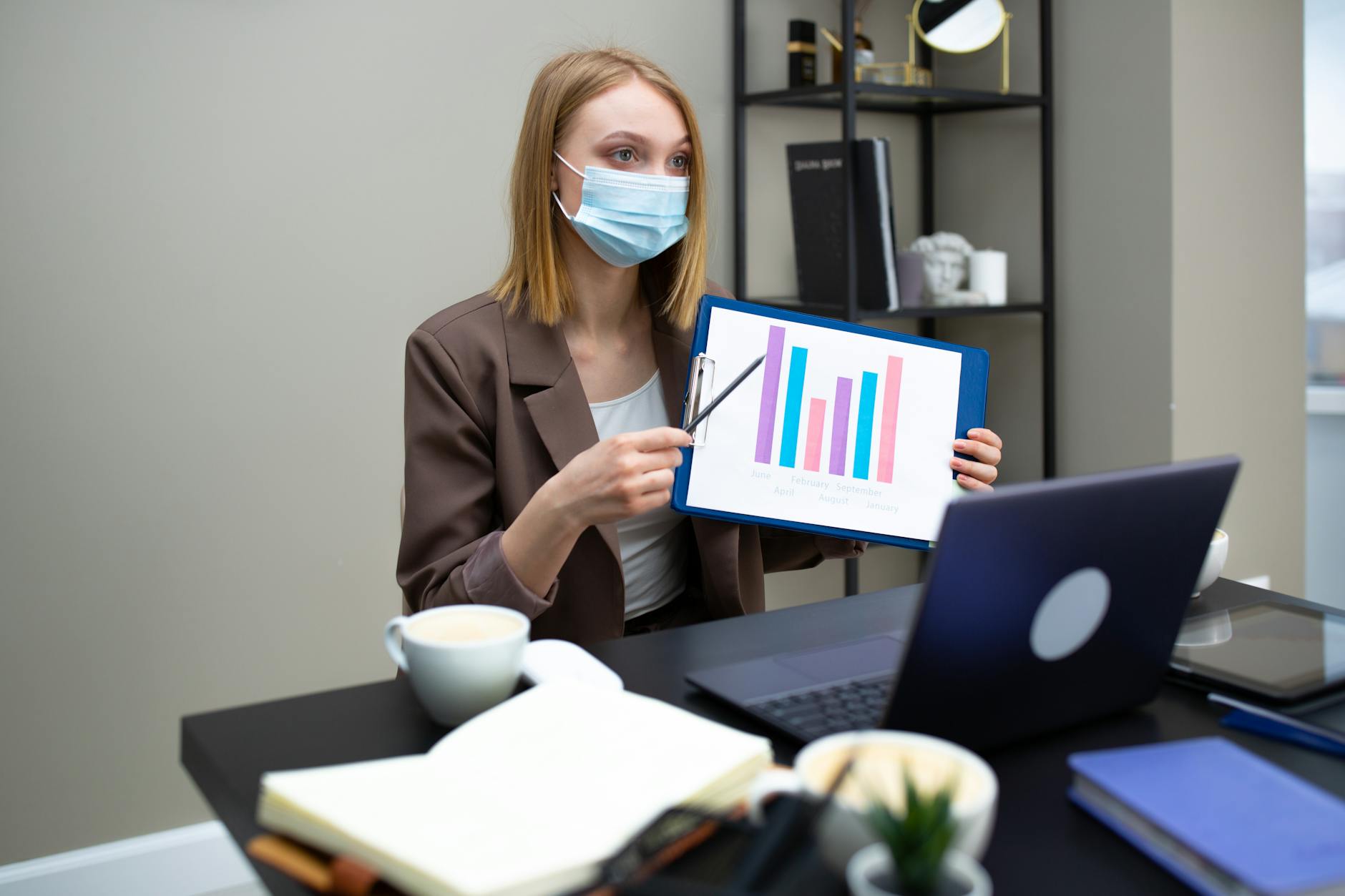 Professional woman wearing mask showing data in an office environment during a video call, focusing on new normal work practices.
